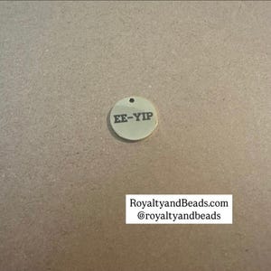 May include: A small, gold-colored, circular charm with the engraved text "EE-YIP" and a small hole at the top. The charm is set against a neutral, tan background. The text "RoyaltyandBeads.com @royaltyandbeads" is displayed below.