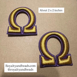 May include: Two embroidered patches in the shape of the Greek letter Omega. Each patch is approximately 2 x 2 inches, featuring gold thread on a purple background. The text "Royaltyandbeads.com @royaltyandbeads" is visible.