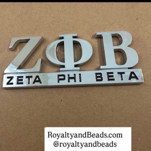 May include: A shiny silver Zeta Phi Beta emblem. The letters Z, Φ, and B are displayed above the words ZETA, PHI, and BETA. The emblem is on a brown surface. The text "RoyaltyandBeads.com @royaltyandbeads" is at the bottom.