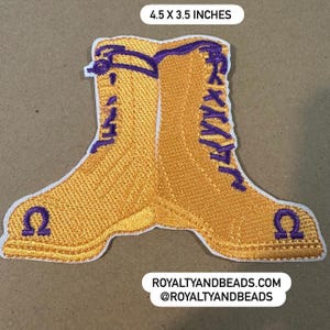 May include: Embroidered patch of two yellow boots with purple laces and the Greek letter Omega. The patch measures 4.5 x 3.5 inches. The text "ROYALTYANDBEADS.COM @ROYALTYANDBEADS" is at the bottom.