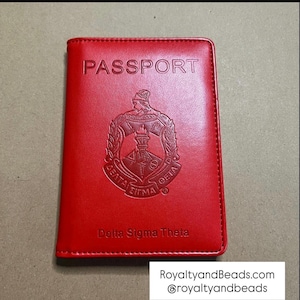 May include: A red passport holder with the word "PASSPORT" embossed at the top. The cover features a Delta Sigma Theta emblem and the text "Delta Sigma Theta" at the bottom.