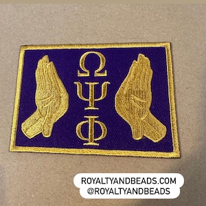 May include: Embroidered patch featuring a purple background with gold trim. The patch displays two gold hands in a praying position, flanking Greek letters. The text "ROYALTYANDBEADS.COM @ROYALTYANDBEADS" is at the bottom.