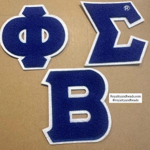 May include: Three blue and white embroidered Greek letter patches. The letters are in a bold, block style with a white border. The letters are Phi, Sigma, and Beta. The image includes the text "Royaltyandbeads.com @royaltyandbeads".