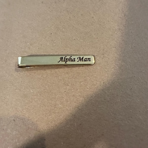 May include: A gold-colored tie clip with the words "Alpha Man" engraved in a cursive font. The clip is rectangular with rounded edges and a hinged clasp.