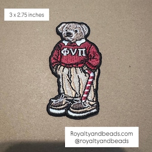 May include: Embroidered patch of a teddy bear wearing a red sweater with white collar and Greek letters, tan pants, and brown shoes. The bear holds a red and white striped candy cane. Patch measures 3 x 2.75 inches.