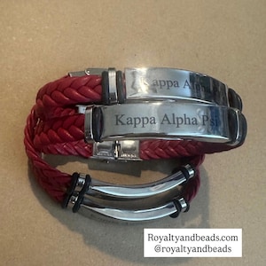 May include: Red braided leather bracelet with silver-tone metal accents. The bracelet features a clasp closure and engraved metal plates with the text "Kappa Alpha Psi". The bracelet is a stylish accessory.