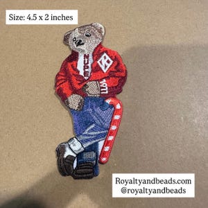 May include: Embroidered patch featuring a bear wearing a red jacket with white lettering and a diamond emblem. The bear is also wearing blue jeans and holding a red and white candy cane. The patch measures 4.5 x 2 inches.