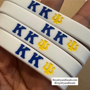 May include: Three white silicone wristbands featuring the Greek letters Kappa Kappa Psi in raised, glossy lettering. The two K letters are colored in a deep royal blue, while the Psi symbol is a bright, vibrant yellow. The bands have a smooth finish.