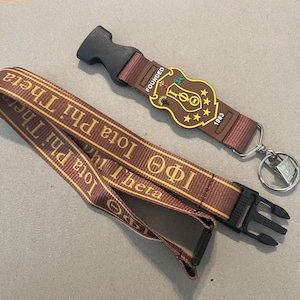 May include: Brown lanyard with gold lettering that reads "Theta Phi Iota" and "1963". The lanyard has a black plastic clip and a silver key ring. A brown rubber charm with gold lettering that reads "Founded 1963" is attached to the lanyard.