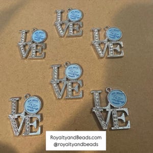 May include: Silver-toned metal charms spelling "LOVE", with clear rhinestone accents on the letters. Each charm has a light blue circular emblem with text. The bottom of the image has the text "RoyaltyandBeads.com" and "@royaltyandbeads".