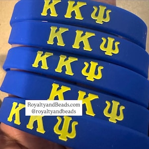 May include: A stack of blue rubber wristbands with yellow lettering. Each band features the Greek letters K, K, and a Psi symbol. The text "RoyaltyandBeads.com @royaltyandbeads" is printed on one of the bands.