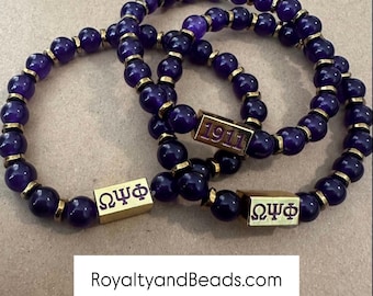 May include: Three beaded bracelets with dark purple beads and gold-colored accents. Each bracelet features a gold-colored rectangular bead with the Greek letters ΩΨΦ or the number 1911. The text "RoyaltyandBeads.com @royaltyandbeads" is at the bottom.