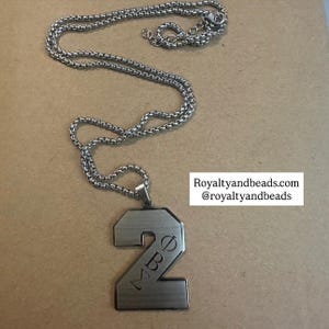 May include: Silver-toned necklace with a pendant shaped like the number 2. The pendant has the letters "MBE" engraved on it. The chain is a linked design. The background is a light brown surface.