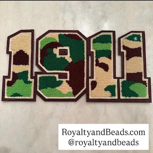 May include: Embroidered patches spelling out "1911" in a collegiate style. The numbers feature a camouflage pattern in shades of green, brown, and tan, outlined with a dark brown border. The text "RoyaltyandBeads.com @royaltyandbeads" is at the bottom.