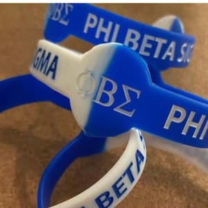 May include: A collection of blue and white silicone wristbands. The bands feature the Greek letters Phi Beta Sigma, with the letters and text in silver. The wristbands are a mix of blue and white, with the text in silver.
