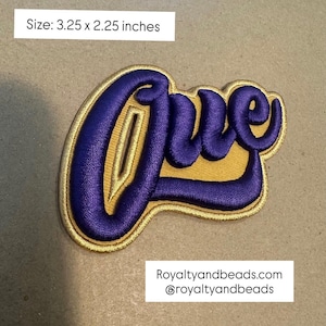 May include: Embroidered patch in purple with gold trim, featuring the word "Que" in a stylized script. The patch measures 3.25 x 2.25 inches. The design is on a tan background.