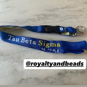 May include: Blue and yellow lanyard with the text "Tau Beta Sigma" and the Instagram handle "@royaltyandbeads".