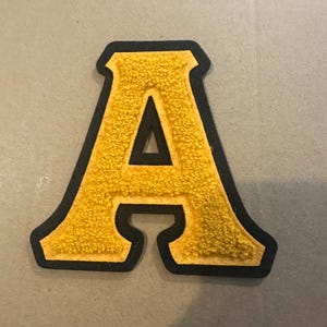 May include: Yellow chenille letter "A" patch with a black border. The letter has a textured, fuzzy appearance. The patch is likely for a jacket or bag.