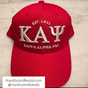 May include: A red baseball cap with white embroidered text. The text includes "EST. 1911", "KAY", and "KAPPA ALPHA PSI". The cap has a curved brim and a button on top.