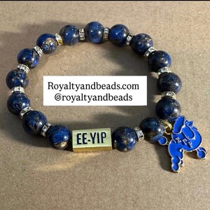 May include: A dark blue beaded bracelet with gold accents and a blue poodle charm. The bracelet features large, round beads with a marbled effect, interspersed with smaller, rhinestone-studded beads. A gold rectangular bead reads "EE-YIP". The text "Royaltyandbeads.com @royaltyandbeads" is also visible.