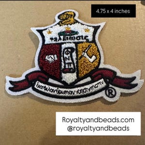 May include: Embroidered patch, 4.75 x 4 inches, featuring a shield design with burgundy, gold, and white sections. The patch includes Greek lettering and a banner with additional text. The patch has a white border and a burgundy ribbon.