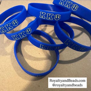 May include: A collection of blue rubber wristbands with the Greek letters "KKΨ" in white and yellow. The wristbands are scattered, with some overlapping. The text "RoyaltyandBeads.com @royaltyandbeads" is at the bottom.