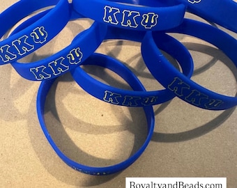 Könnte beinhalten: Eine Ansammlung blauer Gummi-Armbänder mit den griechischen Buchstaben "KKΨ" in Weiß und Gelb. Die Armbänder sind verstreut, einige überlappen sich. Der Text "RoyaltyandBeads.com @royaltyandbeads" ist unten zu sehen.
