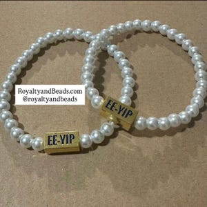 May include: Two white pearl bracelets with gold-colored rectangular beads. The beads have the text "EE-YIP" in blue. The bracelets are on a brown surface. The website RoyaltyandBeads.com is visible in the image.