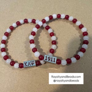 May include: Two beaded bracelets with alternating red and white beads. Each bracelet has a rectangular silver charm with the Greek letters "KAΨ" and the number "1911". The bracelets are displayed on a light brown surface.