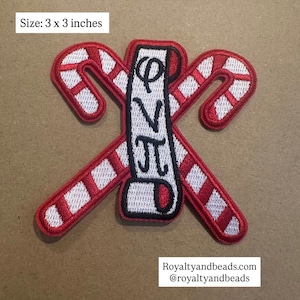 May include: A 3 x 3 inch embroidered patch featuring two red and white candy canes crossed with a vertical white banner. The banner has black Greek letters. The patch has a black border.