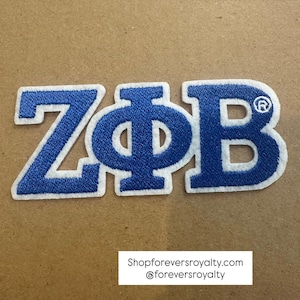 May include: A blue and white embroidered patch featuring the Greek letters Z, Φ, and B. The letters are in a bold, sans-serif font with a white border. The patch is on a tan background. The text "Shopforeversroyalty.com @foreversroyalty" is at the bottom.