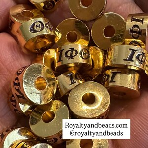May include: Close-up of gold-colored metal beads with black lettering. The beads are cylindrical with a hole in the center. Some beads have the Greek letters I, Φ, and Θ. The website address Royaltyandbeads.com and the Instagram handle @royaltyandbeads are visible.