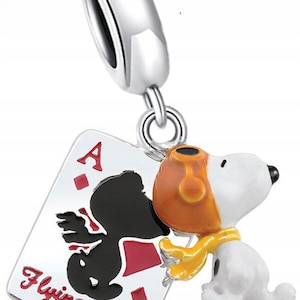 Peut inclure: Un charm argenté représentant Snoopy en tant qu'As Volant, avec un corps blanc, un casque orange et une écharpe jaune. Le charm est suspendu à une boucle argentée et est attaché à une carte à jouer avec l'As de carreau et les mots "Flying Ace".