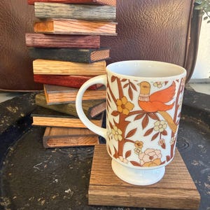 Puede incluir: Una taza de cerámica blanca con base de pedestal presenta un patrón repetitivo de un pájaro naranja y flores. Junto a la taza hay una pila vertical de diez posavasos rectangulares de madera en varios tonos naturales.