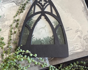 May include: Black gothic-style window frame with a forest scene inside. The frame is placed on a book with a map design. A string of green, round-leafed plants is in the foreground.