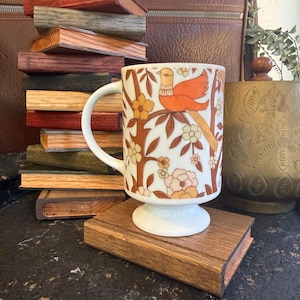 May include: A white ceramic mug with a pedestal base featuring a repeating pattern of orange birds perched on brown branches surrounded by cream and orange flowers. The mug has a curved handle and is positioned on a rectangular wooden block.