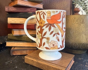 May include: A white ceramic mug with a pedestal base featuring a repeating pattern of orange birds perched on brown branches surrounded by cream and orange flowers. The mug has a curved handle and is positioned on a rectangular wooden block.