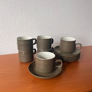 May include: A set of six brown ceramic coffee cups and saucers. The cups and saucers have a textured, woven pattern. The cups are stacked in groups of two and three, with the saucers arranged nearby. The interior of the cups is white.
