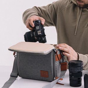 Könnte beinhalten: Eine graue Kameratasche mit beigem oberem Klappdeckel, grauem Schultergurt und braunen Lederakzenten. Eine Person legt eine schwarze Kamera in die Tasche. Zusätzliche Kameraobjektive und eine zweite Kamera sind sichtbar.