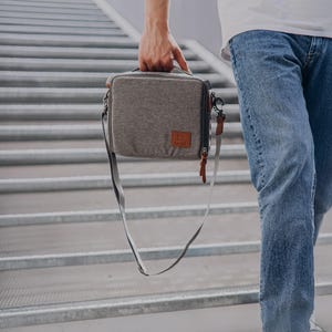 May include: A grey, rectangular bag with a shoulder strap and a handle. The bag features a brown leather patch with a logo and a brown leather zip pull. The bag is being carried by a person wearing blue jeans.