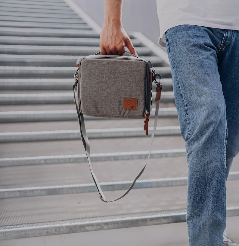 May include: A grey, rectangular bag with a shoulder strap and a handle. The bag features a brown leather patch with a logo and a brown leather zip pull. The bag is being carried by a person wearing blue jeans.