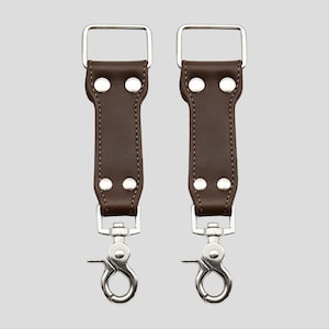 May include: Two brown leather strap connectors with silver-colored metal hardware. Each connector has a rectangular buckle at the top, two rivets, and a swivel clasp at the bottom. The background is a solid light gray.