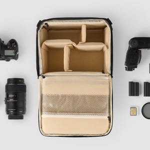 May include: Black camera bag with a beige interior, open to show compartments. Includes a DSLR camera, lenses, flash, memory cards, batteries, and a filter, arranged on a white surface. For photographers.