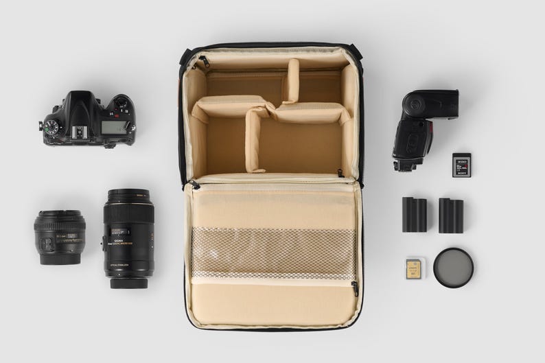 May include: Black camera bag with a beige interior, open to show compartments. Includes a DSLR camera, lenses, flash, memory cards, batteries, and a filter, arranged on a white surface. For photographers.