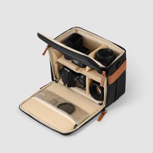 May include: Black camera bag with tan interior compartments, containing a camera, lenses, and a flash. The bag features a brown leather accent and a zipped front pocket. The bag is open, displaying the contents.