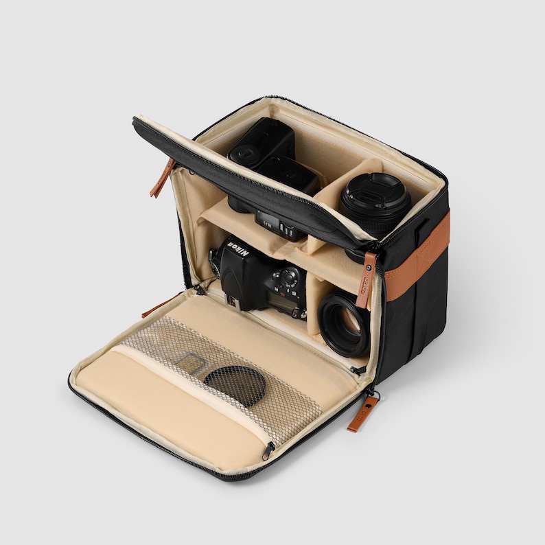 May include: Black camera bag with tan interior compartments, containing a camera, lenses, and a flash. The bag features a brown leather accent and a zipped front pocket. The bag is open, displaying the contents.