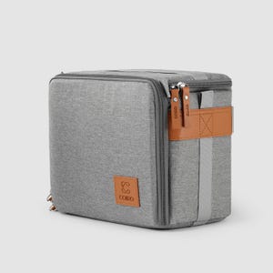 May include: A grey, rectangular cooler bag with a zip closure and brown leather accents. The bag features a brown leather strap and a logo patch. The zips have brown leather pulls with the brand name "COIRO" printed on them.