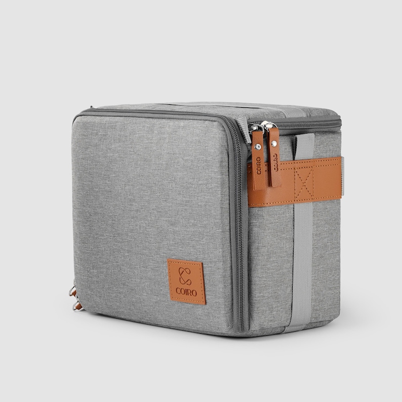 May include: A grey, rectangular cooler bag with a zip closure and brown leather accents. The bag features a brown leather strap and a logo patch. The zips have brown leather pulls with the brand name "COIRO" printed on them.