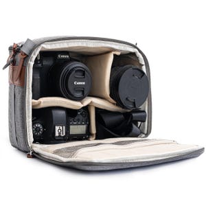May include: Grey camera bag with multiple compartments, containing a Canon camera, lenses, and a flash. The bag features a brown leather accent and a shoulder strap. The interior is beige, and the bag is open, showing the contents.