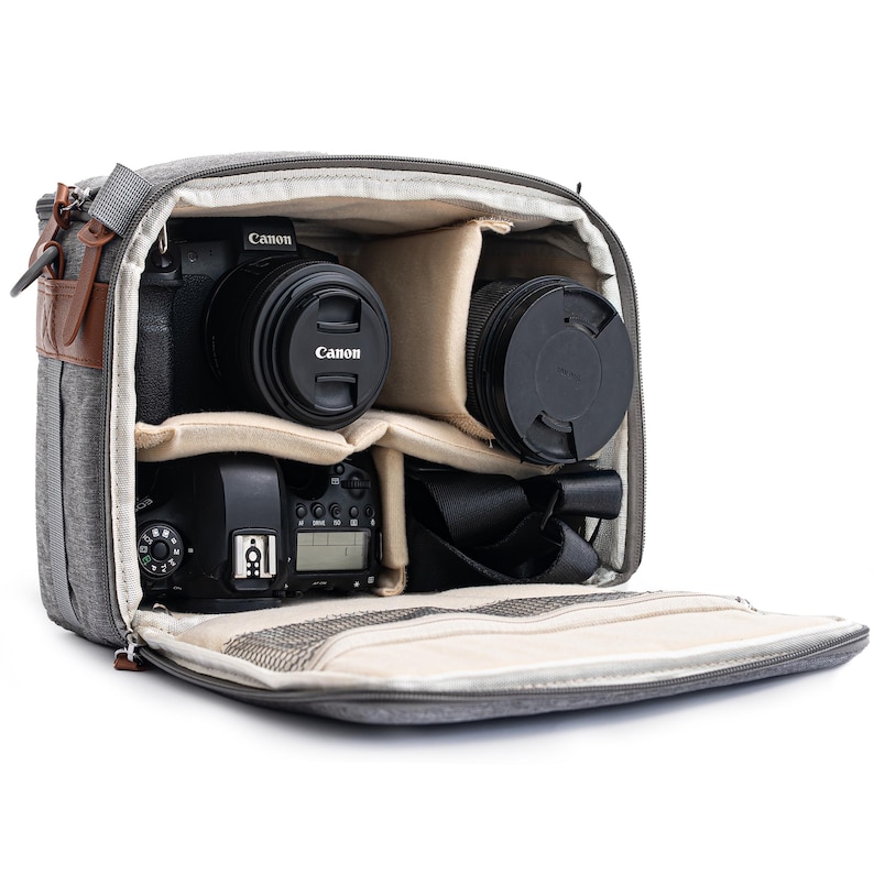 May include: Grey camera bag with multiple compartments, containing a Canon camera, lenses, and a flash. The bag features a brown leather accent and a shoulder strap. The interior is beige, and the bag is open, showing the contents.